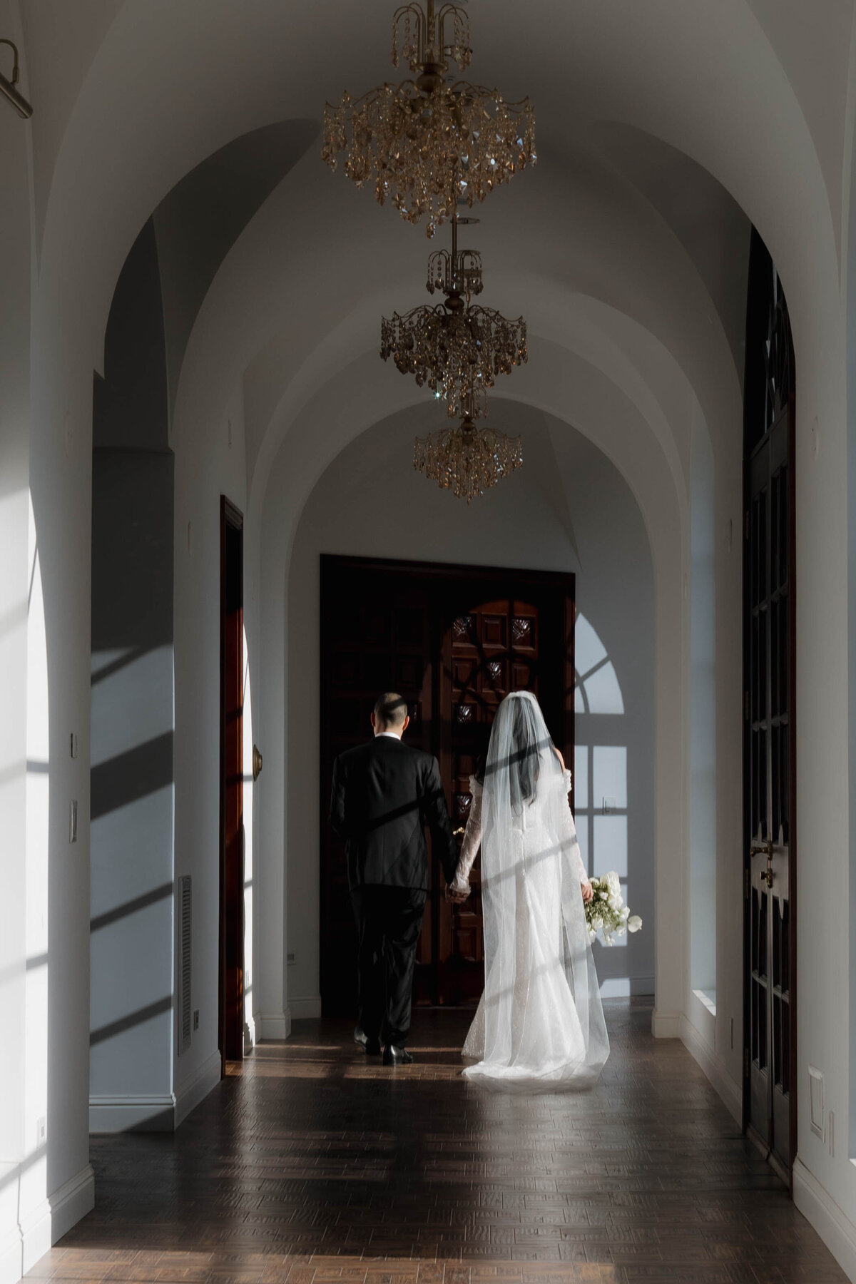 A newlywed couple walks hand in hand through a grand, sunlit hallway with chandeliers, capturing the essence of cinematic wedding photography and understated luxury.