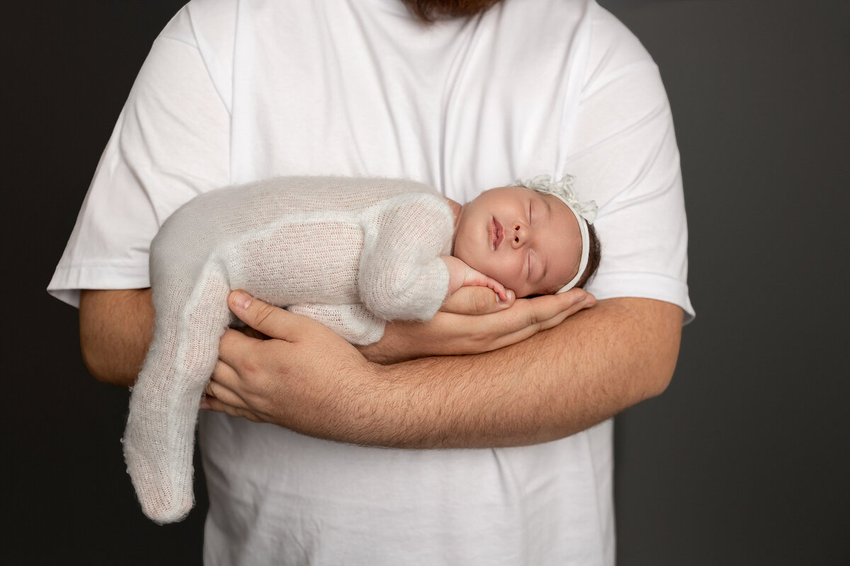 Dad gently holding his newborn daughter swaddled in a soft cream knit outfit with a matching headband, cradled in his arms during their studio session. Captured by Inland Empire newborn photographer Kaitlyn Dawn Photography.