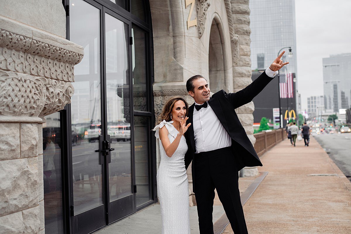 The bride and groom wave to passers by as they stand next to the historic Union Station Hotel in Nashville. The bride is wearing a one shoulder floor length sheath dress with a ruffle over the shoulder. The groom is wearing a black tuxedo with a white shirt and black bow tie. He has one arm around the bride's shoulders and the other is raise up waving hello.