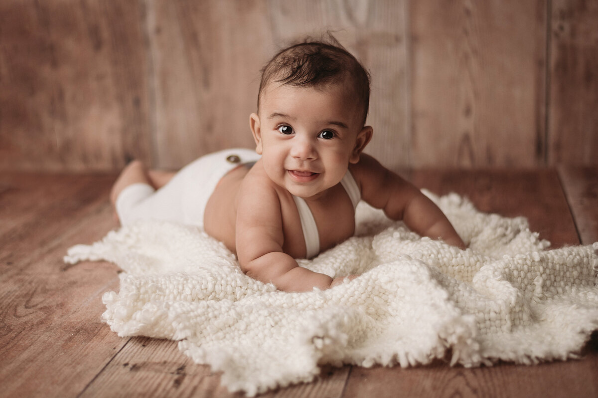 smiling six month old baby boy laying on white fabric over wooden floor laying on tummy in studio