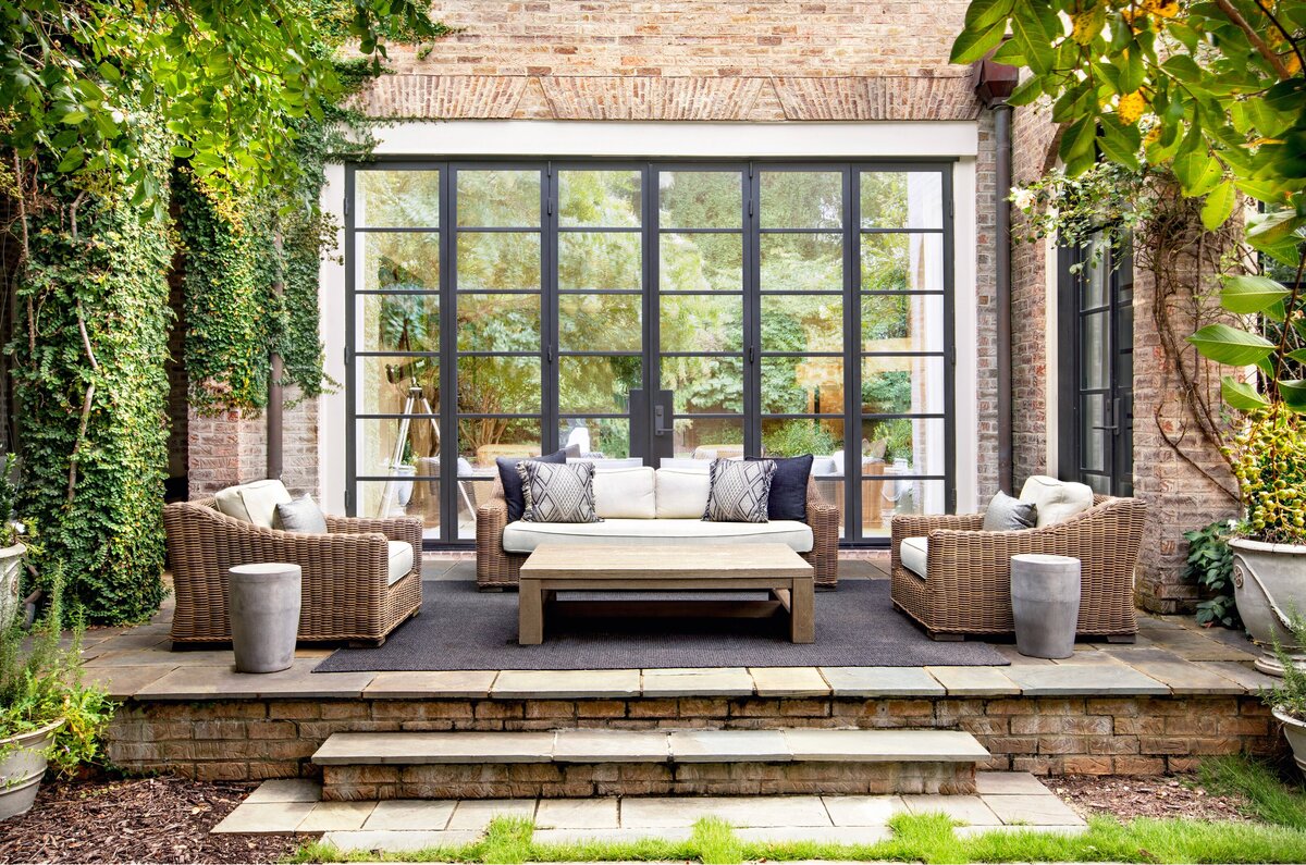 Stylish outdoor patio with wicker furniture, neutral cushions, black-frame glass doors, and lush greenery surrounding the space.