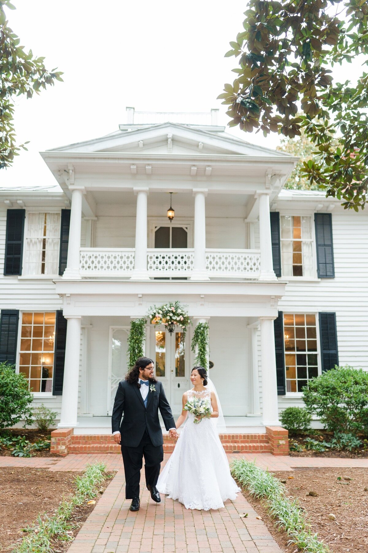 A couple looks at one another and walks hand in hand in front of the Leslie Alford Mims House in Raleigh, NC.