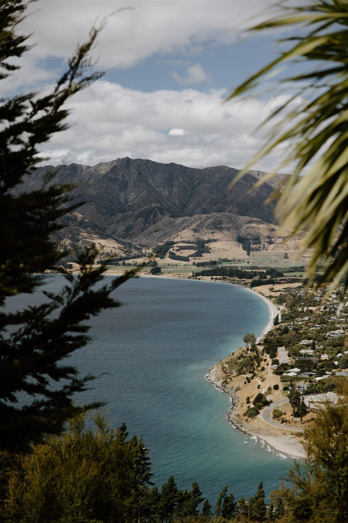 Scenic aerial view of Lake Hawea with turquoise water curving around a bay, framed by mountains and native trees.

