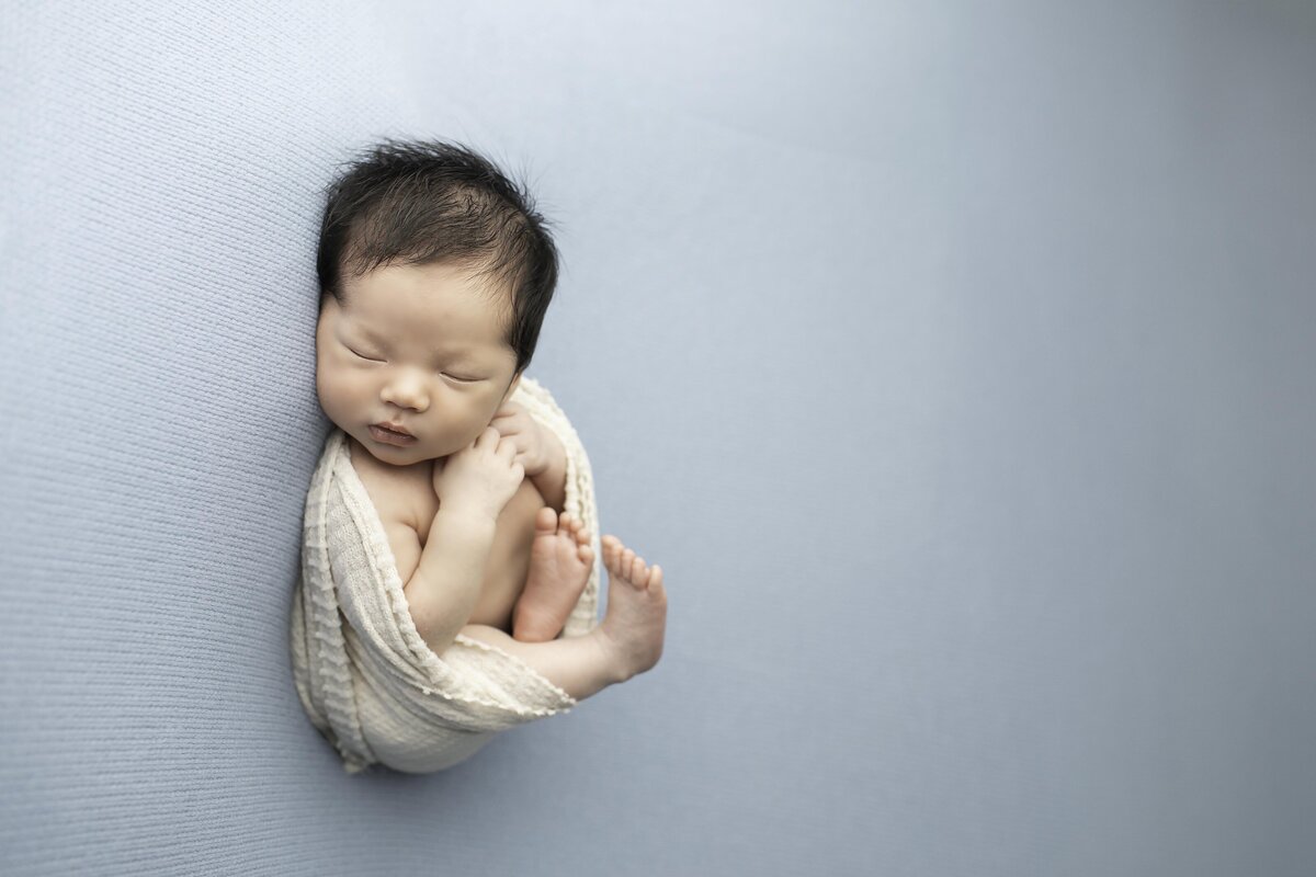 Newborn baby boy with a full head of dark hair, snugly wrapped in a white swaddle, peacefully resting on a soft blue backdrop