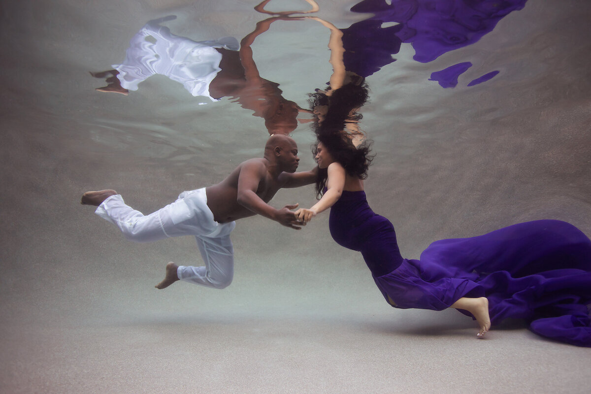 A beautiful underwater maternity photoshoot featuring a couple gracefully reaching for each other, their reflections mirroring their connection. The woman wears a flowing purple gown, showcasing the expertise of a talented Dallas underwater maternity photographer.