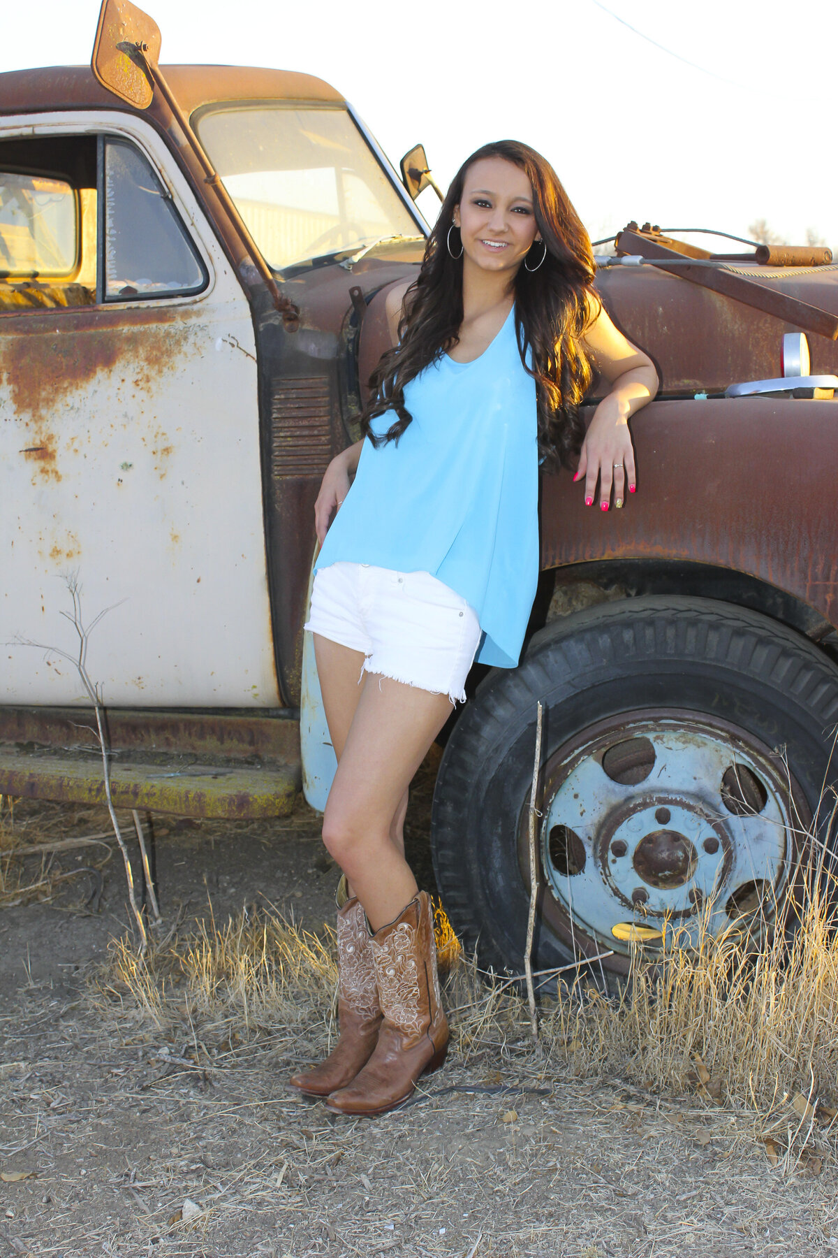 Lady leaning on old rusted truck with glowing light of sunset beaming down golden light