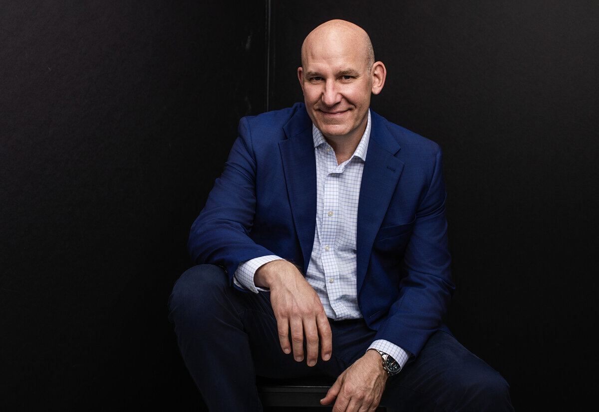 Professional headshot of a confident bald man in a tailored blue suit jacket and checkered dress shirt, seated casually with one arm resting on his knee. Photographed against a dark black studio backdrop at Janel Lee Photography in Cincinnati, Ohio, his approachable smile and relaxed pose convey professionalism and warmth. This headshot highlights the studio’s expertise in creating polished corporate portraits for executives, business leaders, and professionals seeking standout LinkedIn and personal branding images.