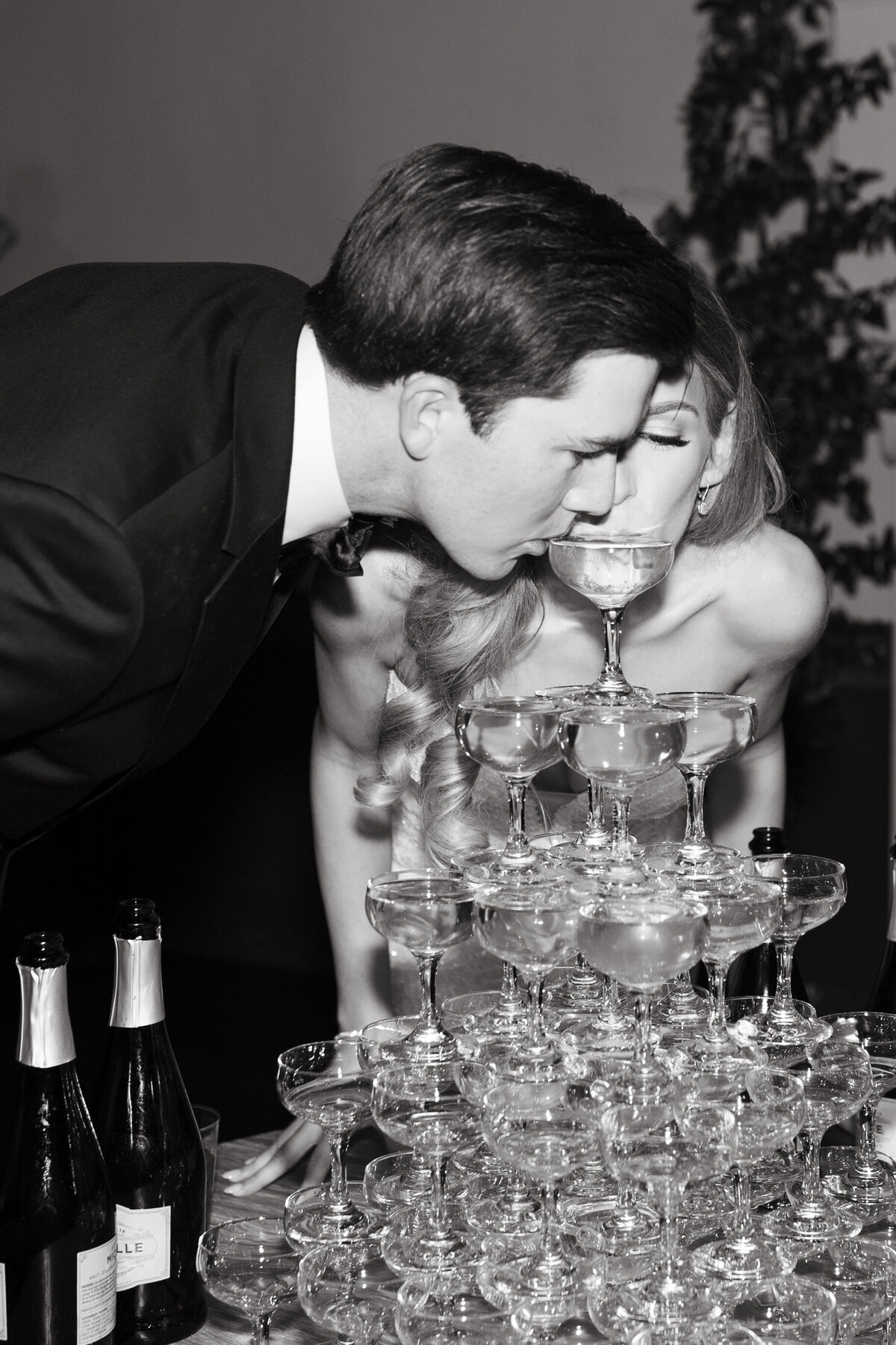 Bride and groom leaning over and sipping from champagne tower at wedding reception