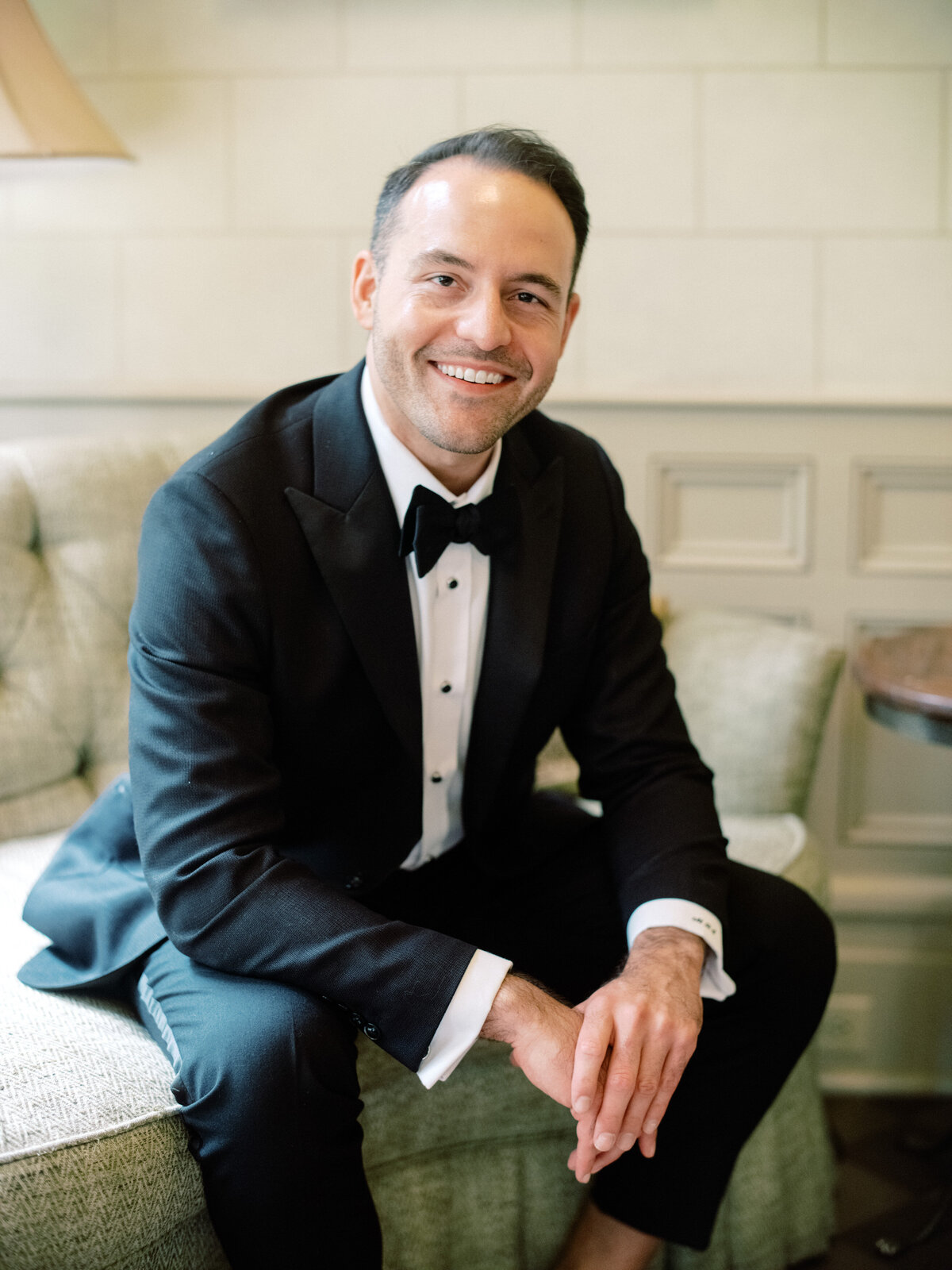 A groom smiling warmly while perched on a couch, showcasing a blend of relaxation and excitement before the wedding ceremony, embodying love and anticipation.