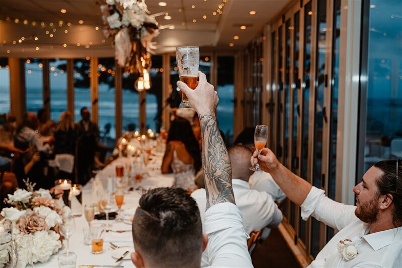 "Gathered in celebration, groomsmen toast to love and laughter at the wedding reception. Join the revelry with this heartwarming glimpse into cherished moments shared among friends. Discover more about the joyous festivities here."