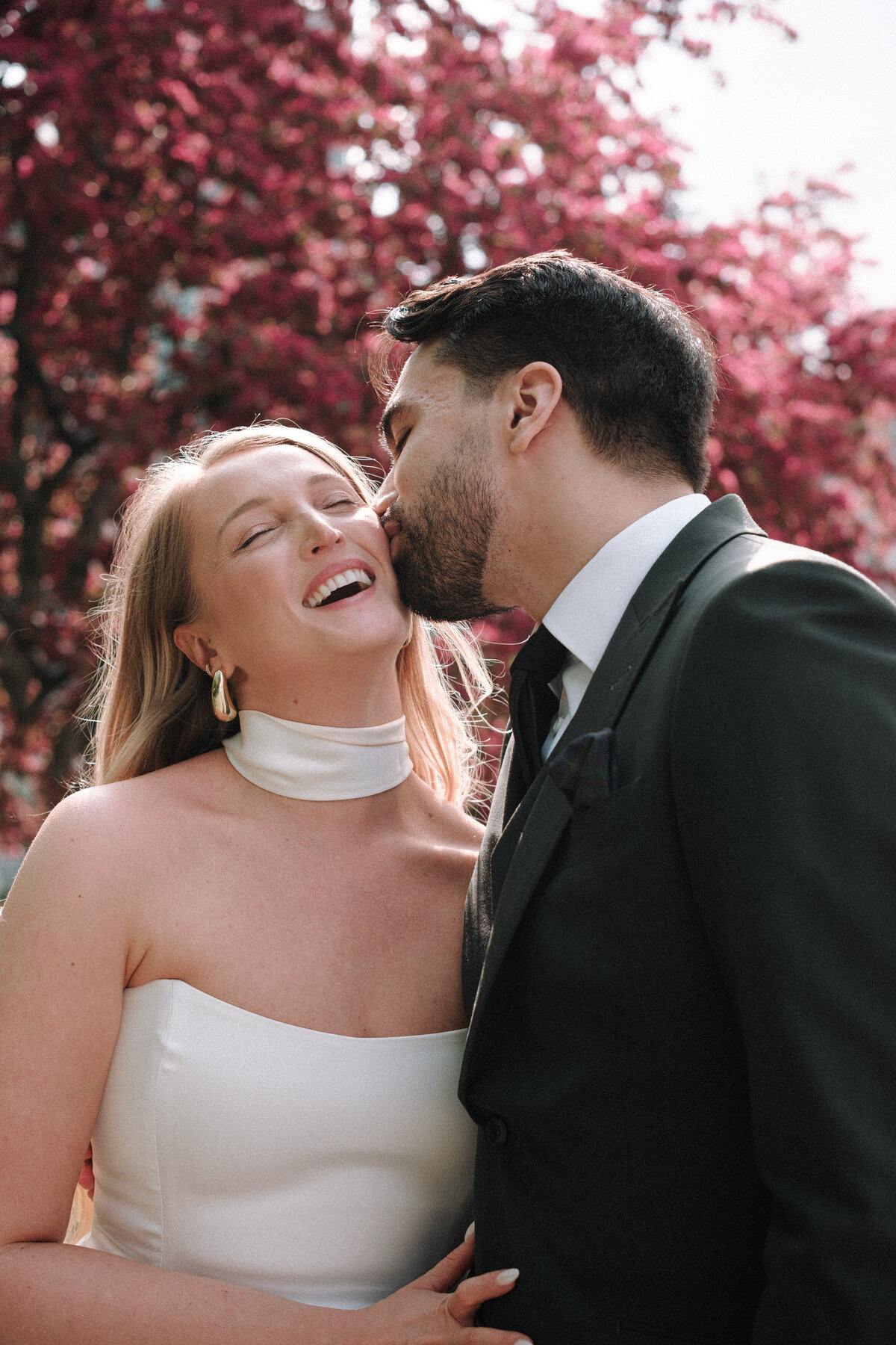 kisses on the cheek from the groom with pink spring flowers behind