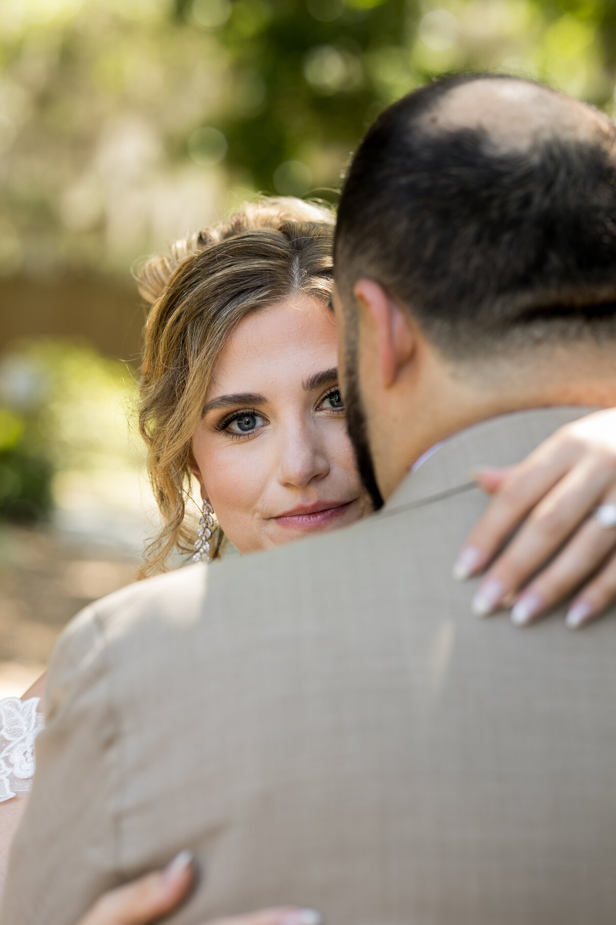  A close-up portrait of a bride embracing her groom during their wedding near the historic Fountain of Youth and along iconic Magnolia Avenue in St. Augustine, Florida. The bride looks softly into the camera over the groom’s shoulder, her hand resting gently on his back, showcasing her elegant engagement ring. Surrounded by soft natural light and the lush backdrop of moss-draped oak trees, this quiet, emotional moment was beautifully captured by Heartful Films, a Jacksonville wedding photographer and videographer known for timeless, heartfelt storytelling.