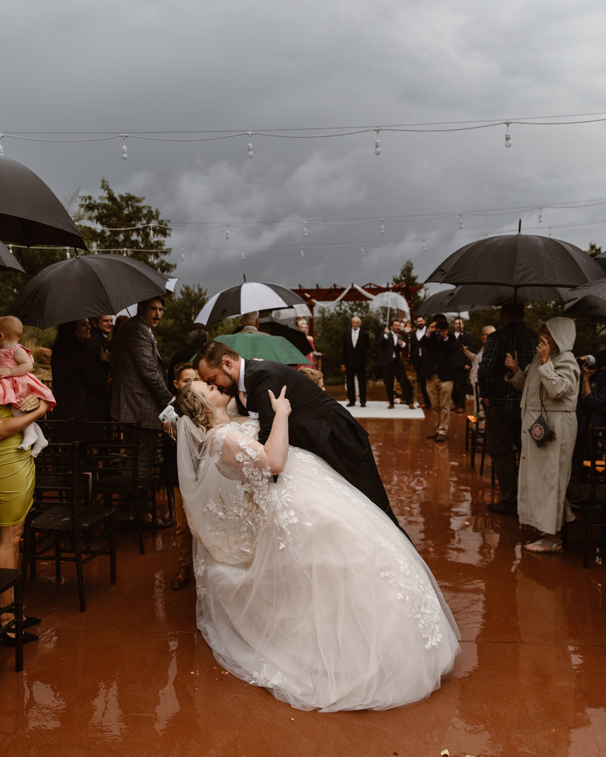 Bride and groom share a dramatic dip kiss during their outdoor wedding ceremony exit, surrounded by guests holding umbrellas under a moody, stormy sky.