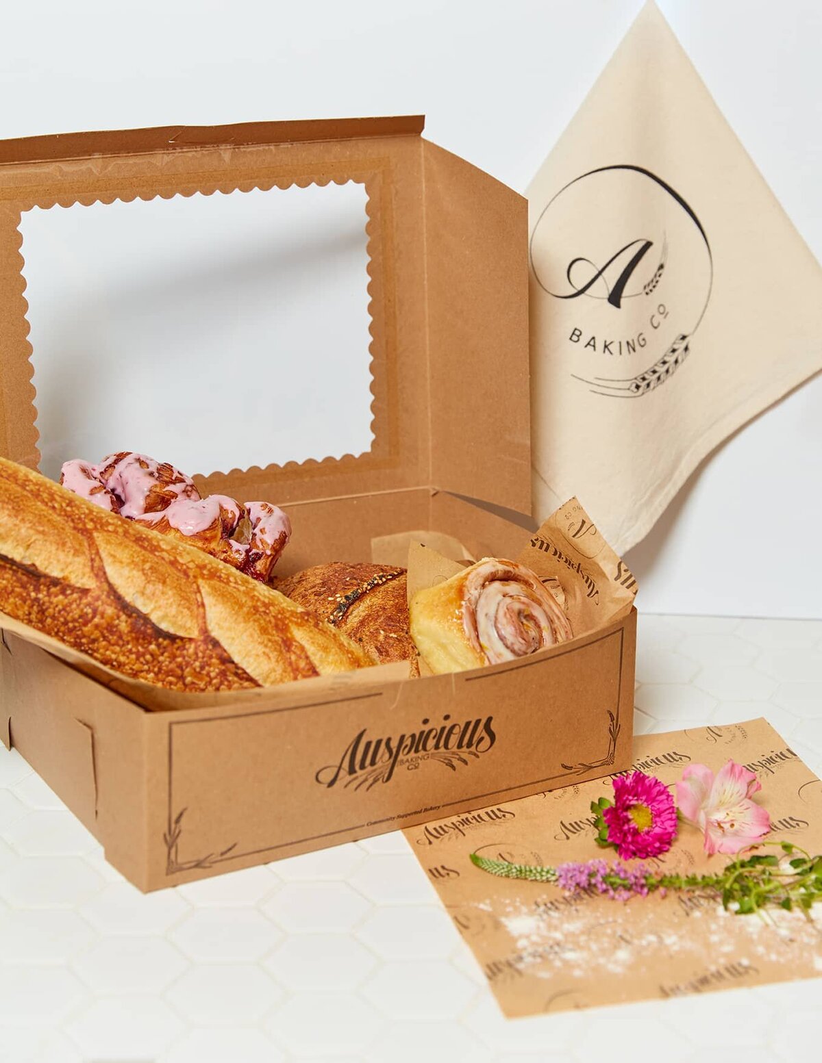 A large box sits open, overflowing with pastries. The front of box is printed with the Auspicious logo and a custom design