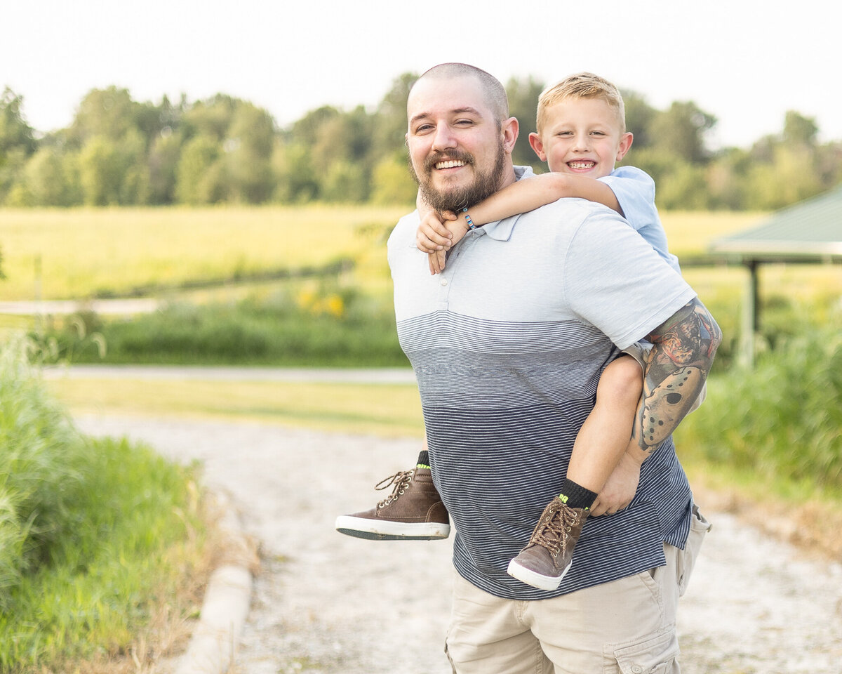Dad and son on a rocked path at a wildlife sanctuary.  Dad has a shaved head with a dark beard, wearing a striped blue shirt, khaki shorts, smiling at the camera with his son on his back.  Son is smiling at the camera with short brown hair, wearing a partial button up short sleeve blue shirt, with khaki shorts, brown shoes, and smiling at the camera