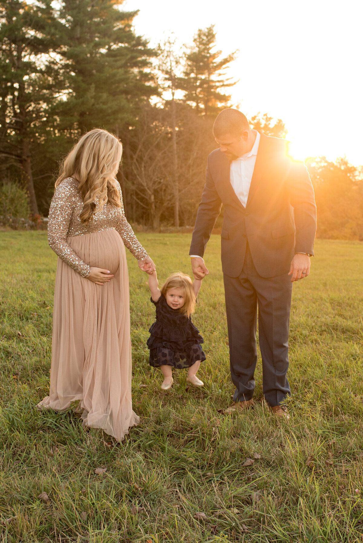 A family of three with a pregnant mom, swinging their daughter in a field at sunset