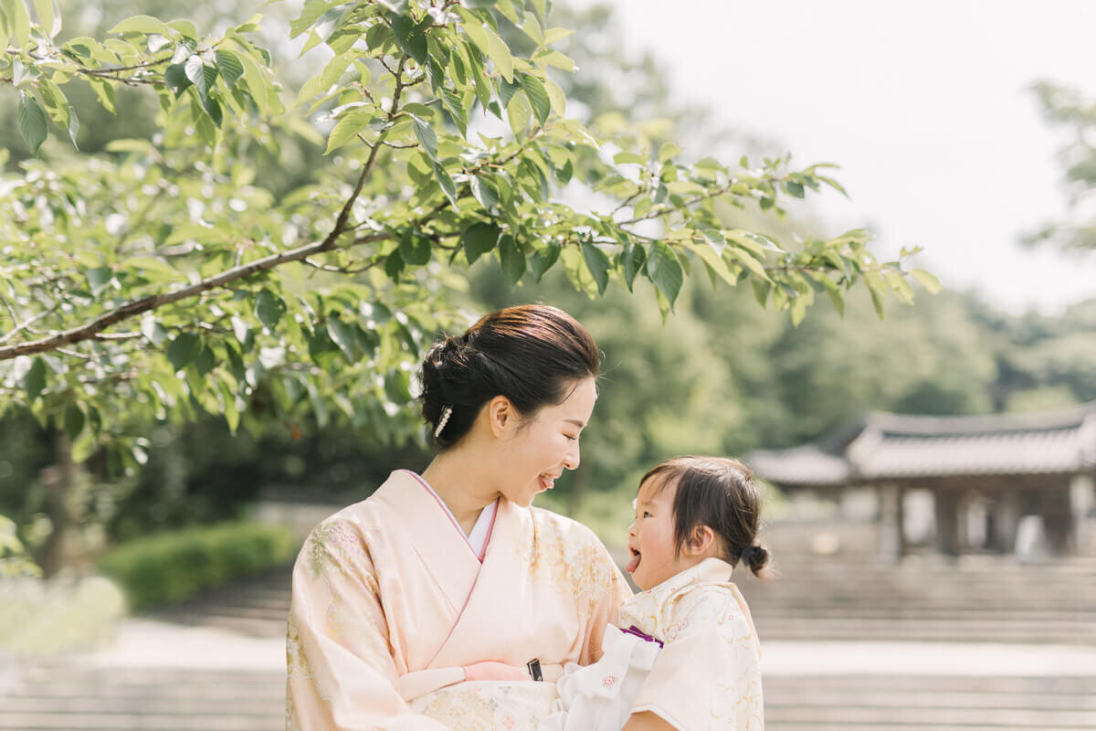 女の子とママが顔を見合わせて、桜の木の下で舌を出して笑っている可愛らしい家族写真。二人とも和装で着物を着ていて正装が素敵な撮影。
