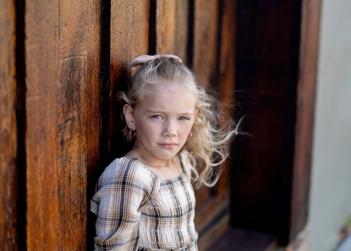 A girl stands alone against a weathered wooden wall for their Iowa Family Photographer