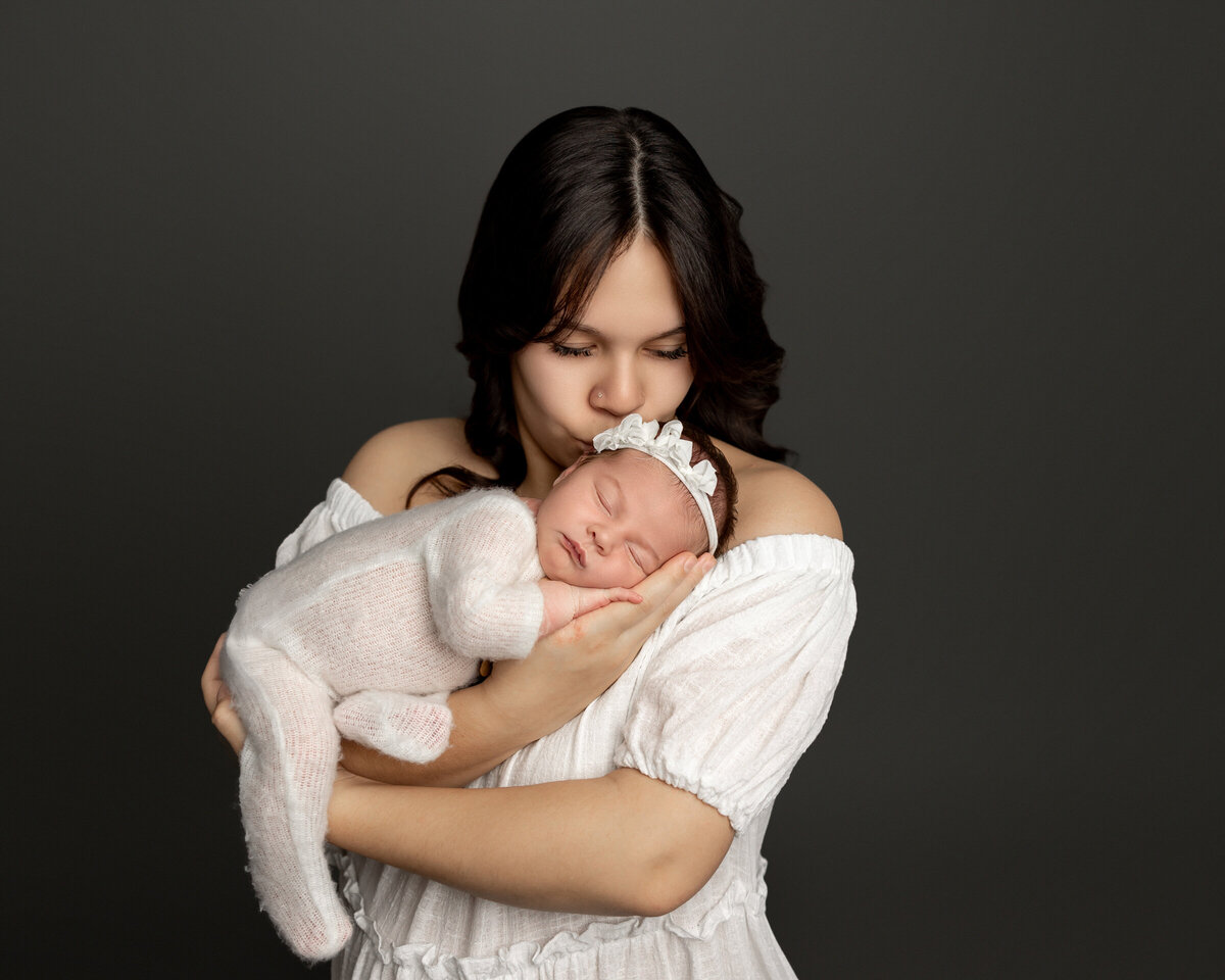 Mom gently kissing her newborn daughter, swaddled in a soft cream knit outfit with a delicate headband, against a dark backdrop during their studio session. Captured by Inland Empire newborn photographer Kaitlyn Dawn Photography.