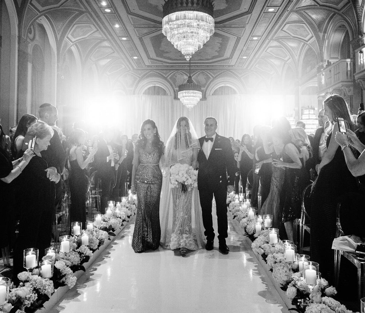 Bride walks down the aisle with her parents during the ceremony black and white