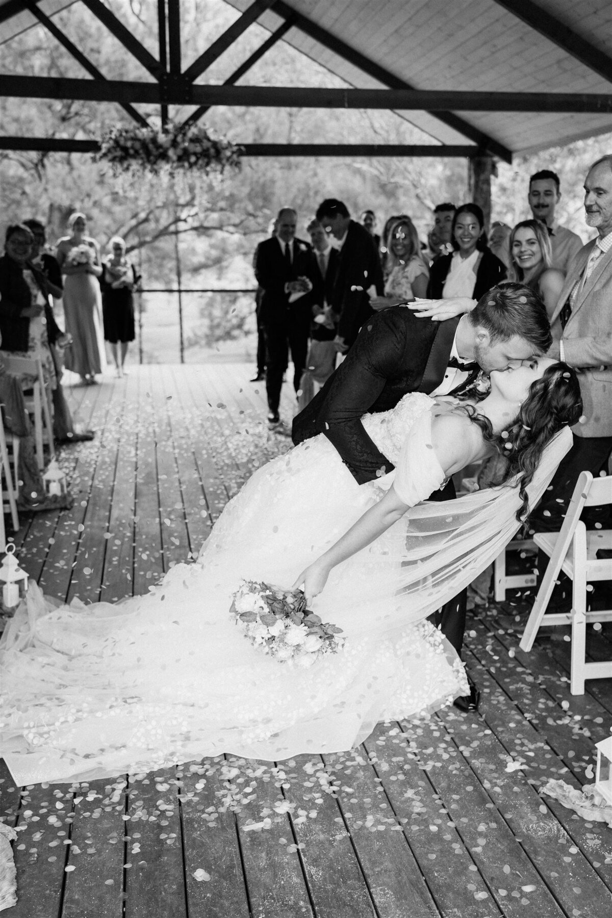 The newlyweds, bathed in golden light, share a tender kiss as they exit the ceremony photoshoot, sealing their love amidst applause and cheers.
