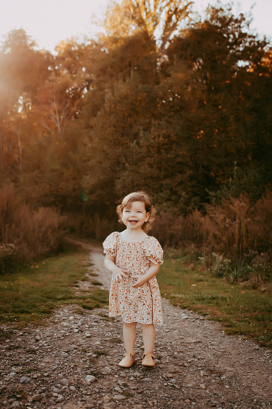 a two year old girl standing on a gravel path smiling in rochester, ny at sunset