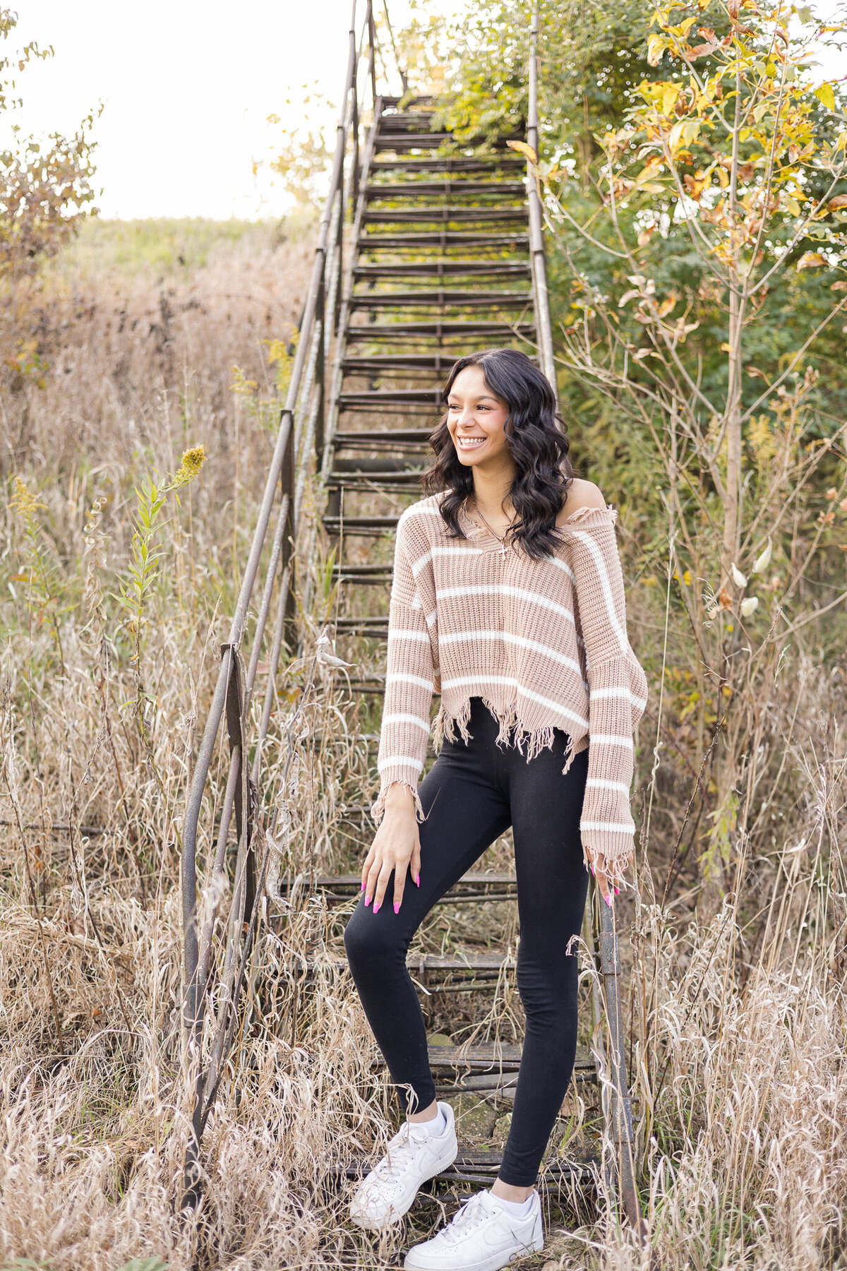 high school female senior, with long black hair, standing in front of a set of stairs, looking away from the camera smiling, wearing black leggings, and light tan and white stripped shirt