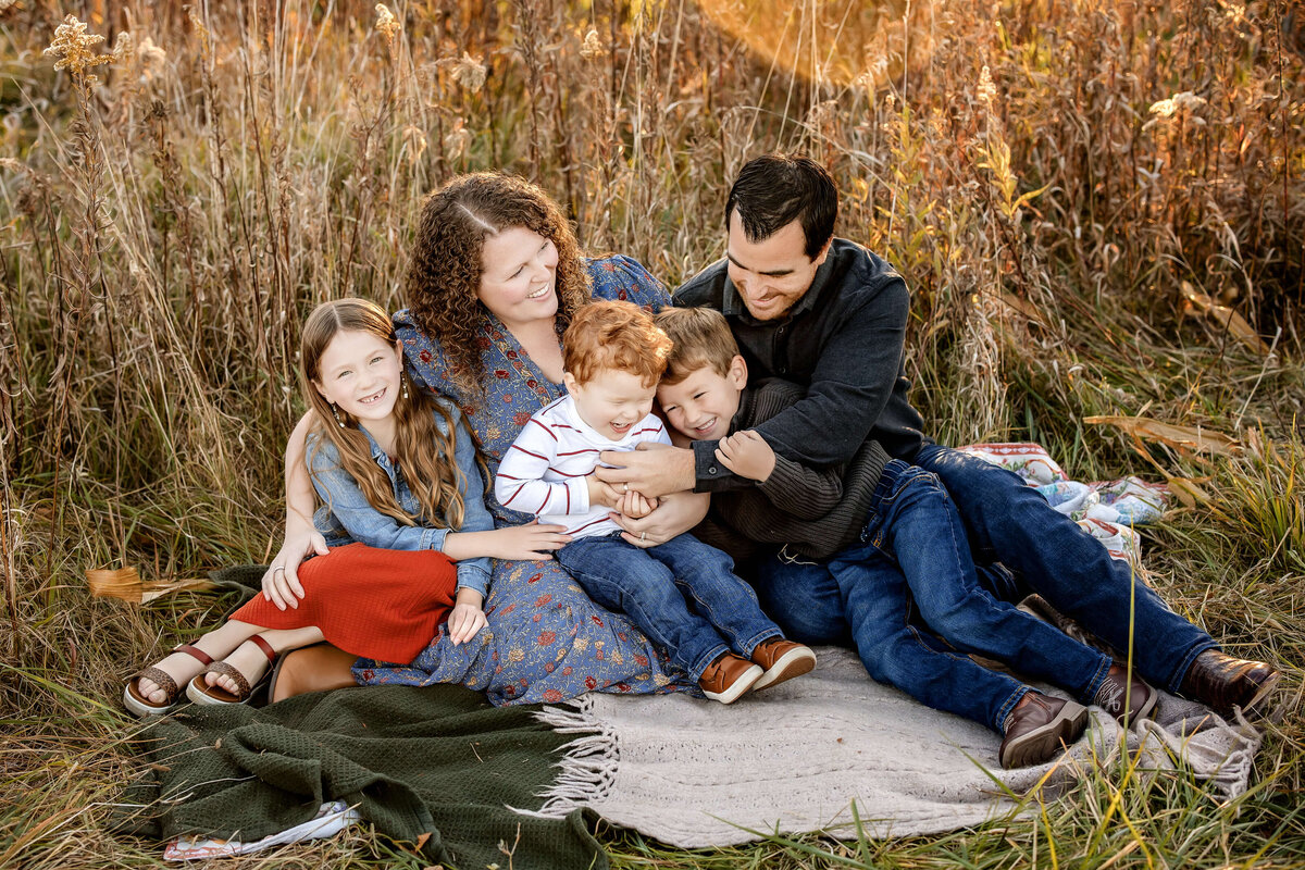 A family of five sits on a blanket in a field. The parents are in the middle, smiling, with their three children around them. The grass is tall and autumnal in color, and everyone appears happy and relaxed.