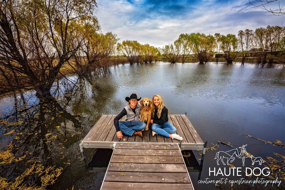 Wide angle view of a couple sitting on a wood dock surrounded by a tree-lined pond with their Golden Retriever.
