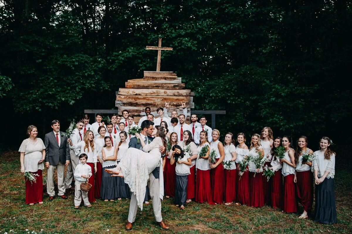 A large wedding group standing in front of a rustic cross, the bride and groom front and center