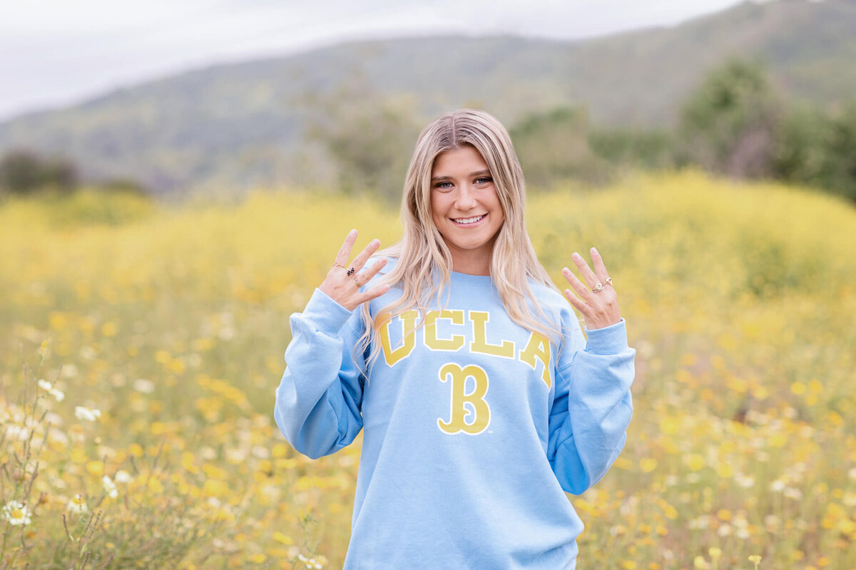 A person with long, blonde hair stands in a field of yellow flowers wearing a light blue UCLA sweatshirt with a large yellow B. The background shows blurred greenery and hills under a cloudy sky. The person is smiling and holding up their fingers.