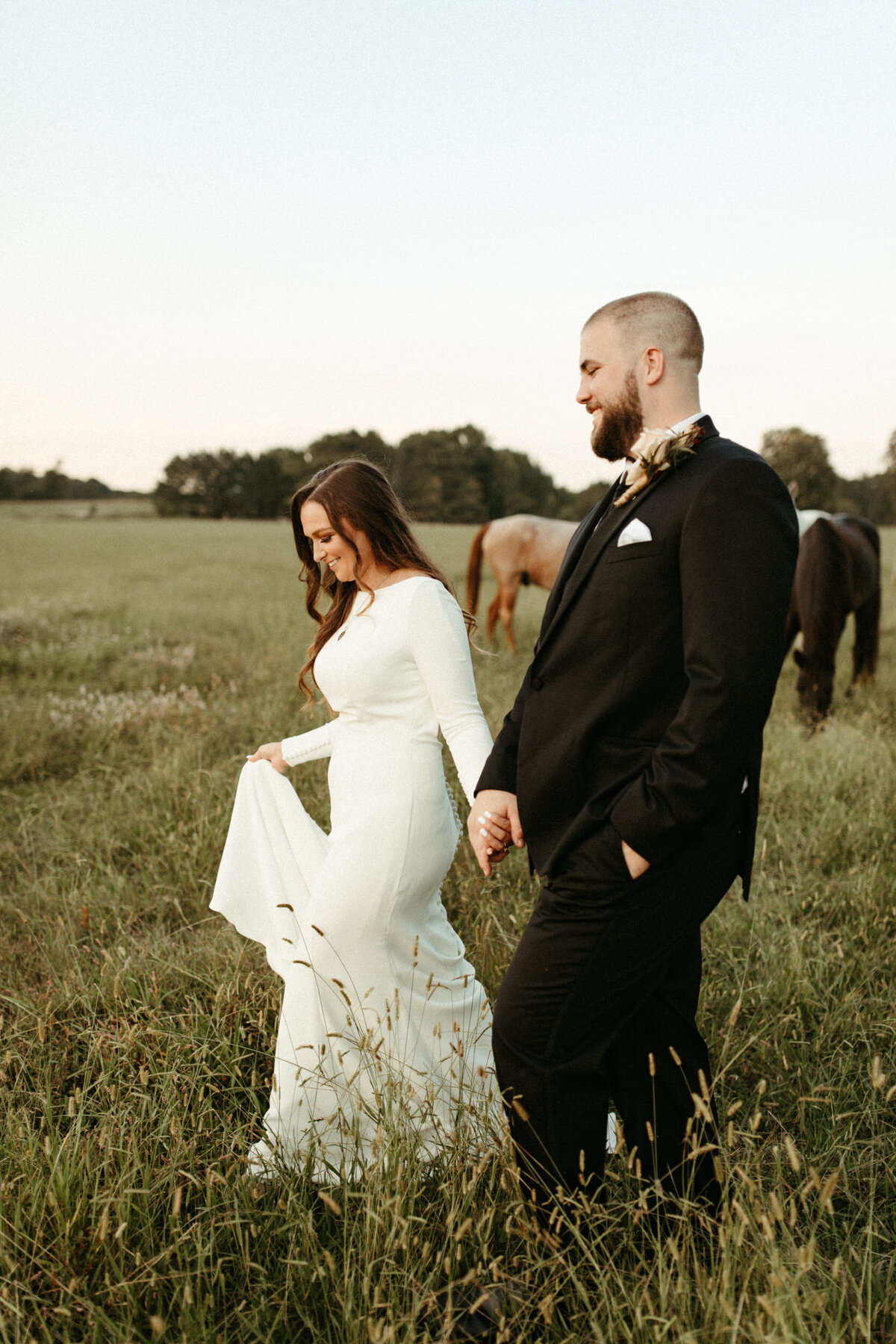 Bride and groom holding hands and walking through a field with horses behind them