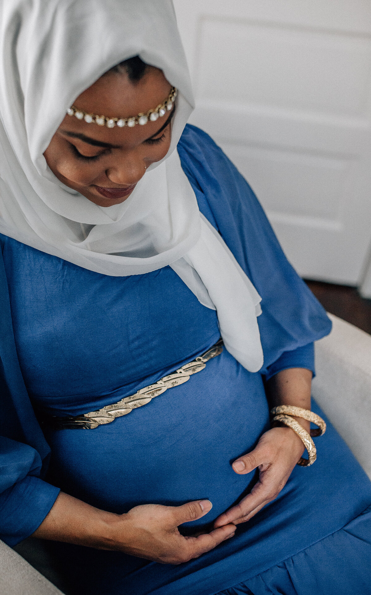 Maternity photoshoot with woman wearing blue dress, white scarf, and holding her belly.