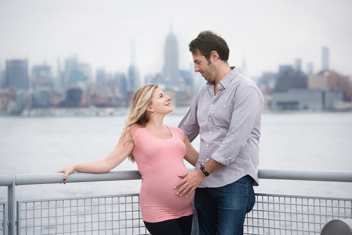 A couple standing on a pier while one places their hand on the other's pregnant stomach