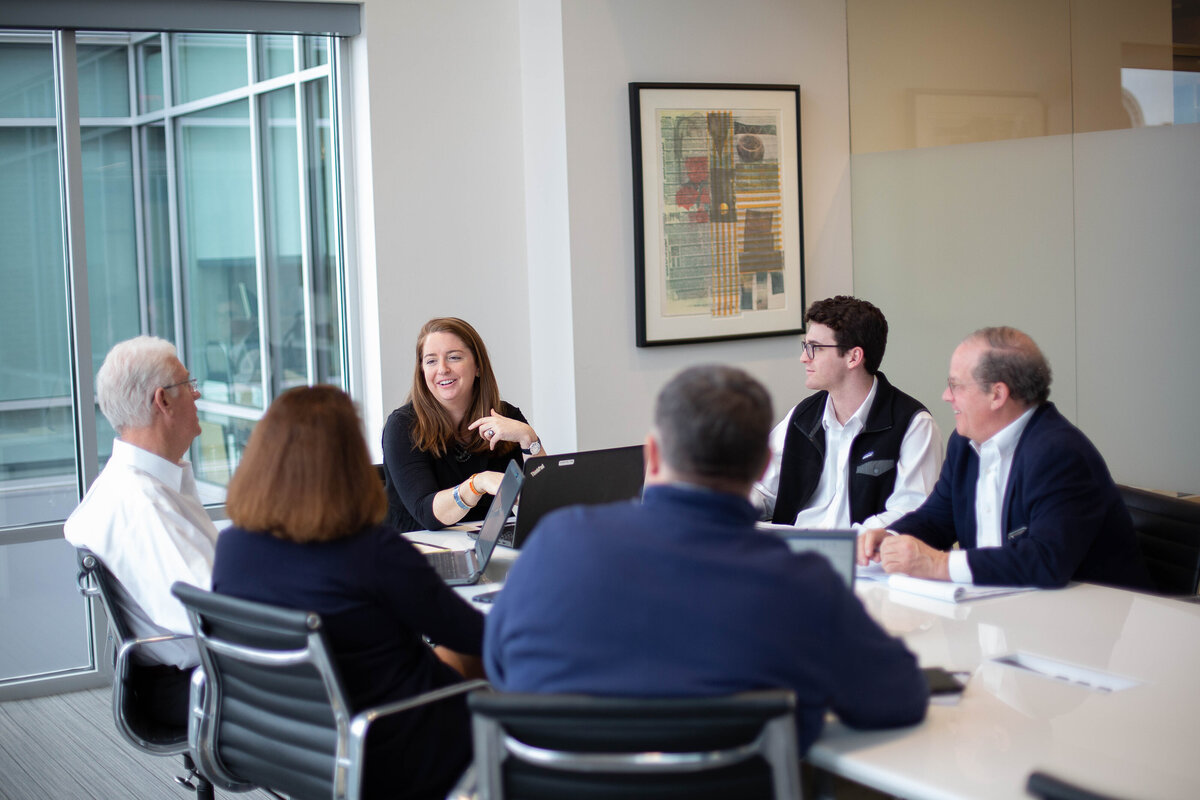 group of businesspeople seated in a glass conference room with laptops open in front of them. A woman is speaking.