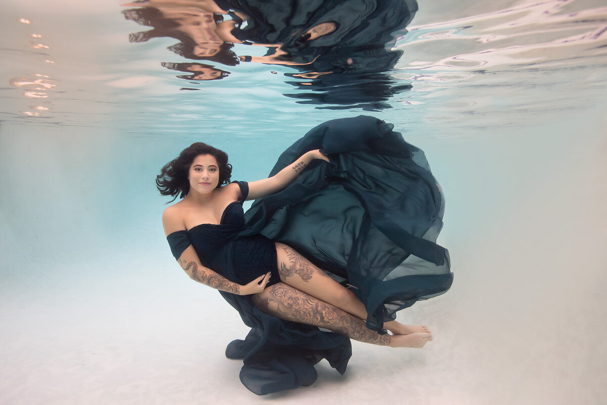 An elegant underwater maternity photo capturing a woman gracefully draped in a flowing dark blue gown, her tattoos beautifully displayed as she floats effortlessly in clear water. The serene expression and soft movements create a dreamy, ethereal look, highlighting the artistry of Dallas underwater maternity photography by Lindsay Walden..