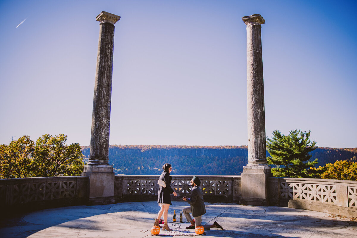 A couple during their proposal with man on his knees proposing in front of pumpkins in the fall at the vista overlook at untermyer gardens in yonkers ny