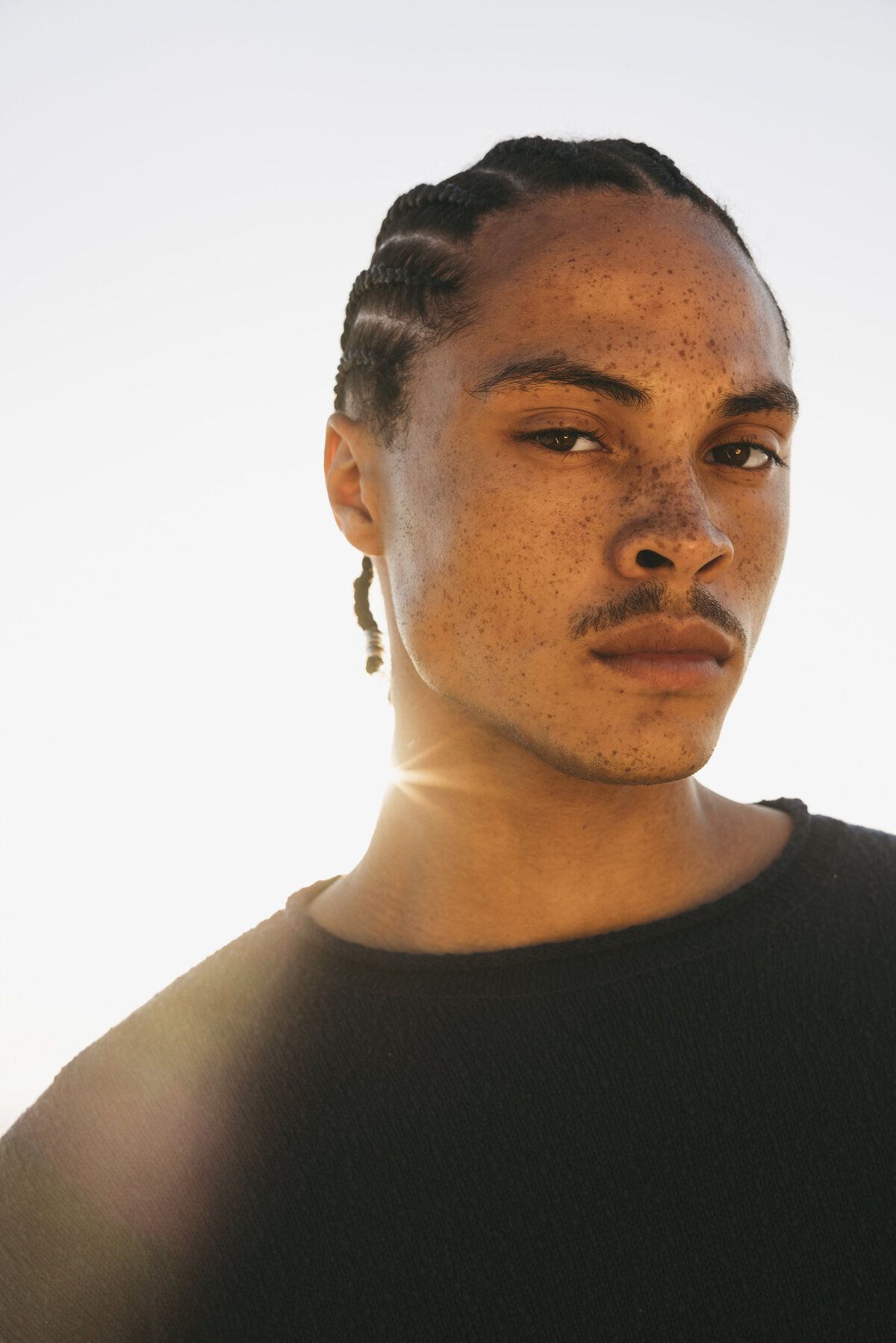 Close-up portrait of Timothe in Cape Town with sunlight flaring over his shoulder, highlighting his freckles and braided hairstyle against a pale sky