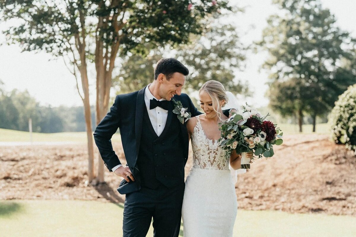 A bride and groom walking arm-in-arm on a golf course, with the bride holding a burgundy and white bouquet, sharing a light-hearted moment.