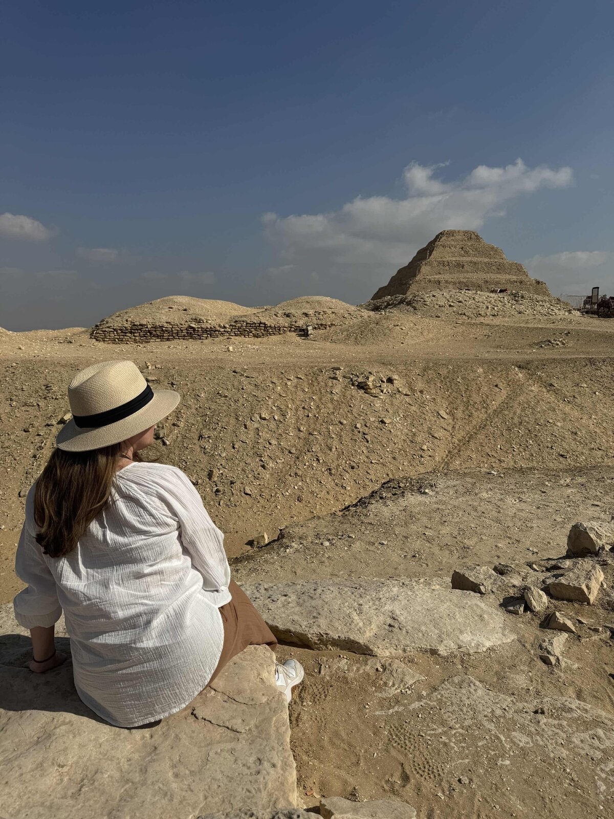 Terra from Adventures by Terra wearing a straw hat and linen clothes sitting on a ledge viewing the Step Pyramid in the distance in Cairo Egypt