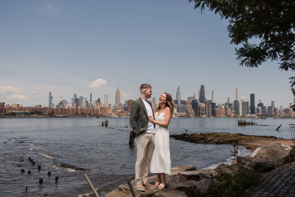 Couple standing on rocks at the East River waterfront in Brooklyn with the Manhattan skyline, including the Empire State Building, in the background; the pair laughs together under a clear summer sky, framed by tree branches and surrounded by remnants of old pilings in the water.