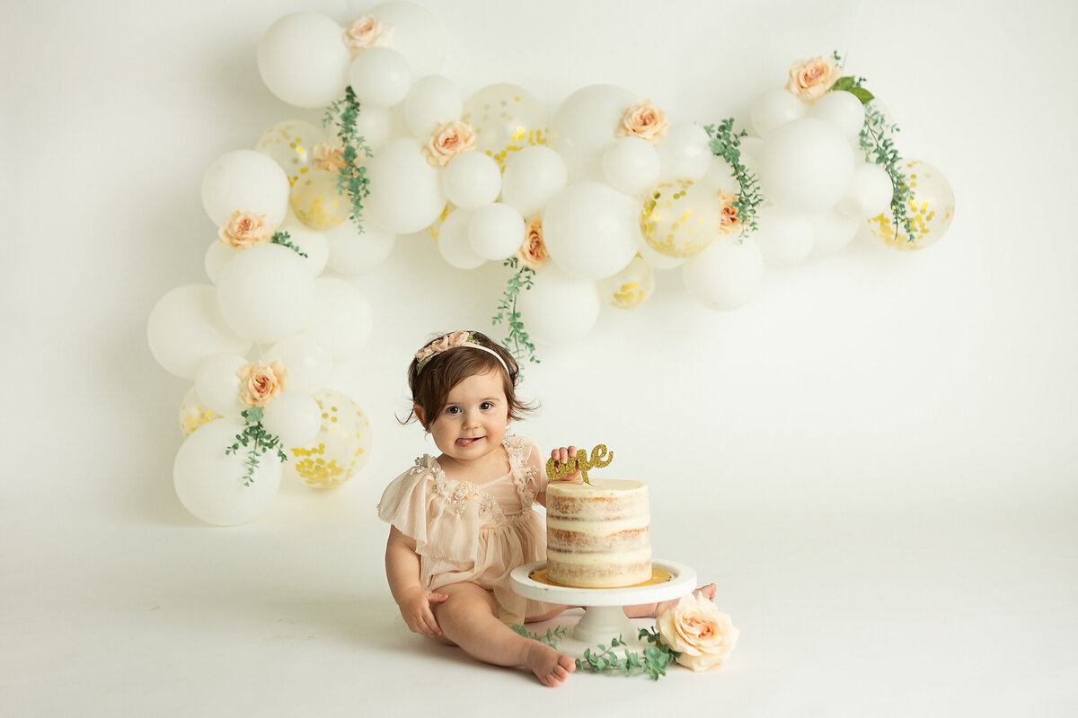 first-birthday-simple-cake-smash-with-white-and-gold-confetti-ballon-garland-simple-florals-baby-girl-in-soft-pink-ruffle-romper