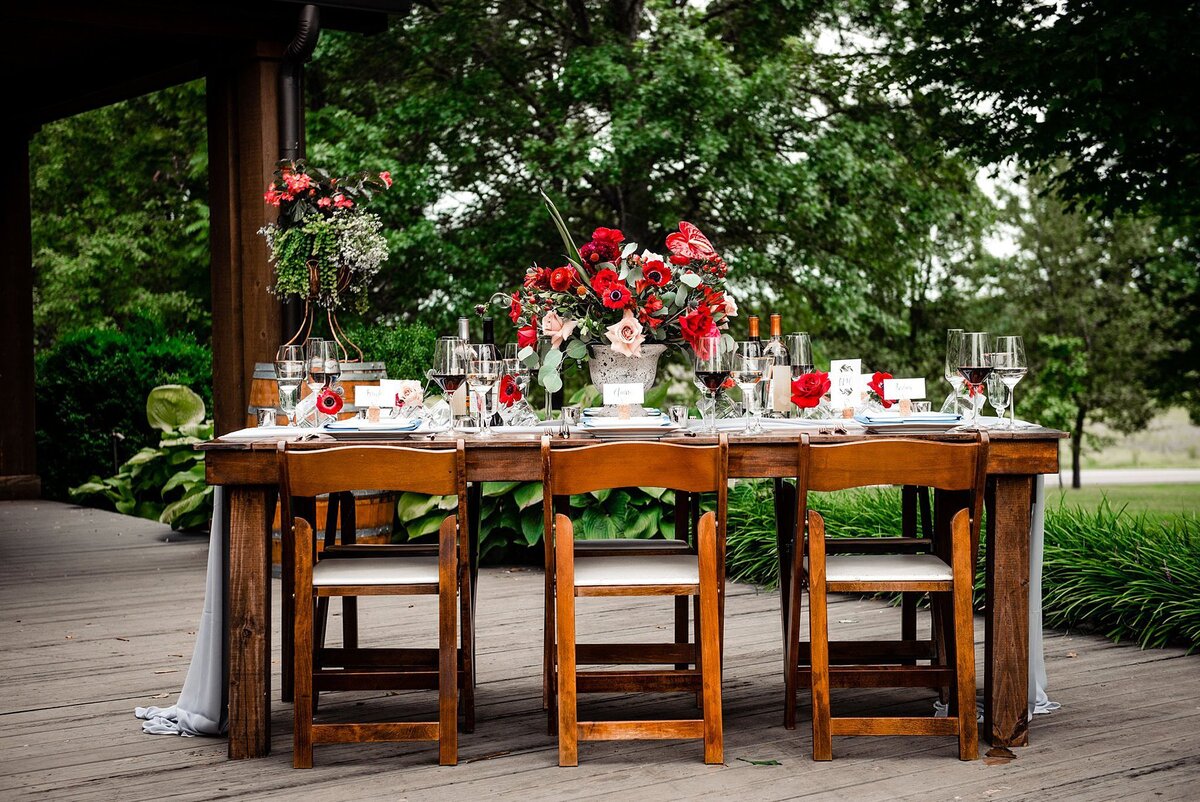 Large dark wood farm table with fruitwood garden chairs on an outdoor patio overlooking a vineyard in Franklin, TN. The table is set with white plates, crystal glassware, silver flatware and a long, sheer dove grey table runner. The large floral centerpiece is bursting with red gerbera daisies, red roses, red peonies, red poppies, and other red flowers. The red bridal bouquet is sitting on a wine barrel in the background at Arrington Vineyard.