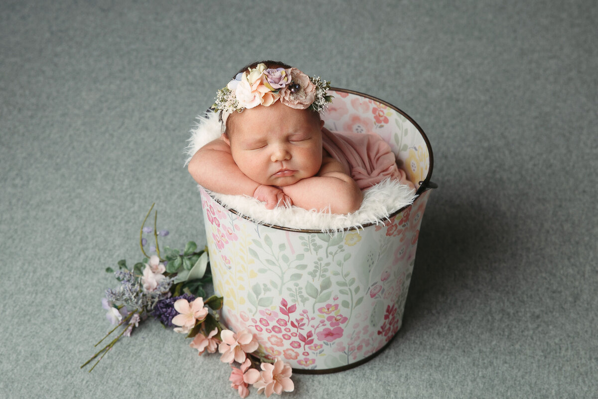 Pretty newborn sitting in a green floral bucket with flower headband and flowers on the floor