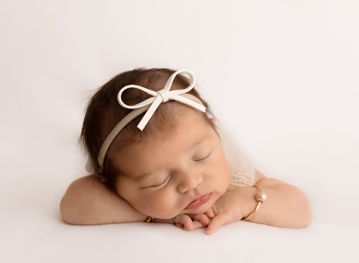 Baby girl is sleeping for her newborn photoshoot. Baby is laying on her belly with her hands folded under her chin. She has a thin bow headband and pearl bracelet. Captured by top Brooklyn newborn photographer Chaya Bornstein