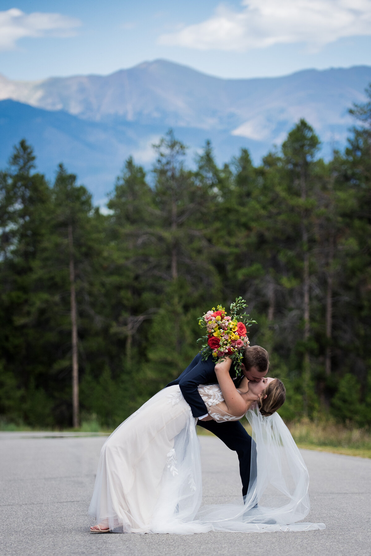 A groom dips his bride for a kiss as she holds her vibrant bouquet with the Colorado mountains in the backkground.