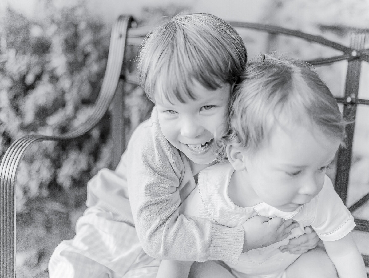 Black and white sibling portrait of two young brothers embracing on a garden bench, capturing their playful bond—photographed by a Birmingham, AL family photographer.