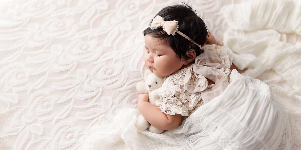 Baby girl wearing a lace outfit and bow headband, lying on her side and cuddling a small plush bear on a soft white fabric backdrop.