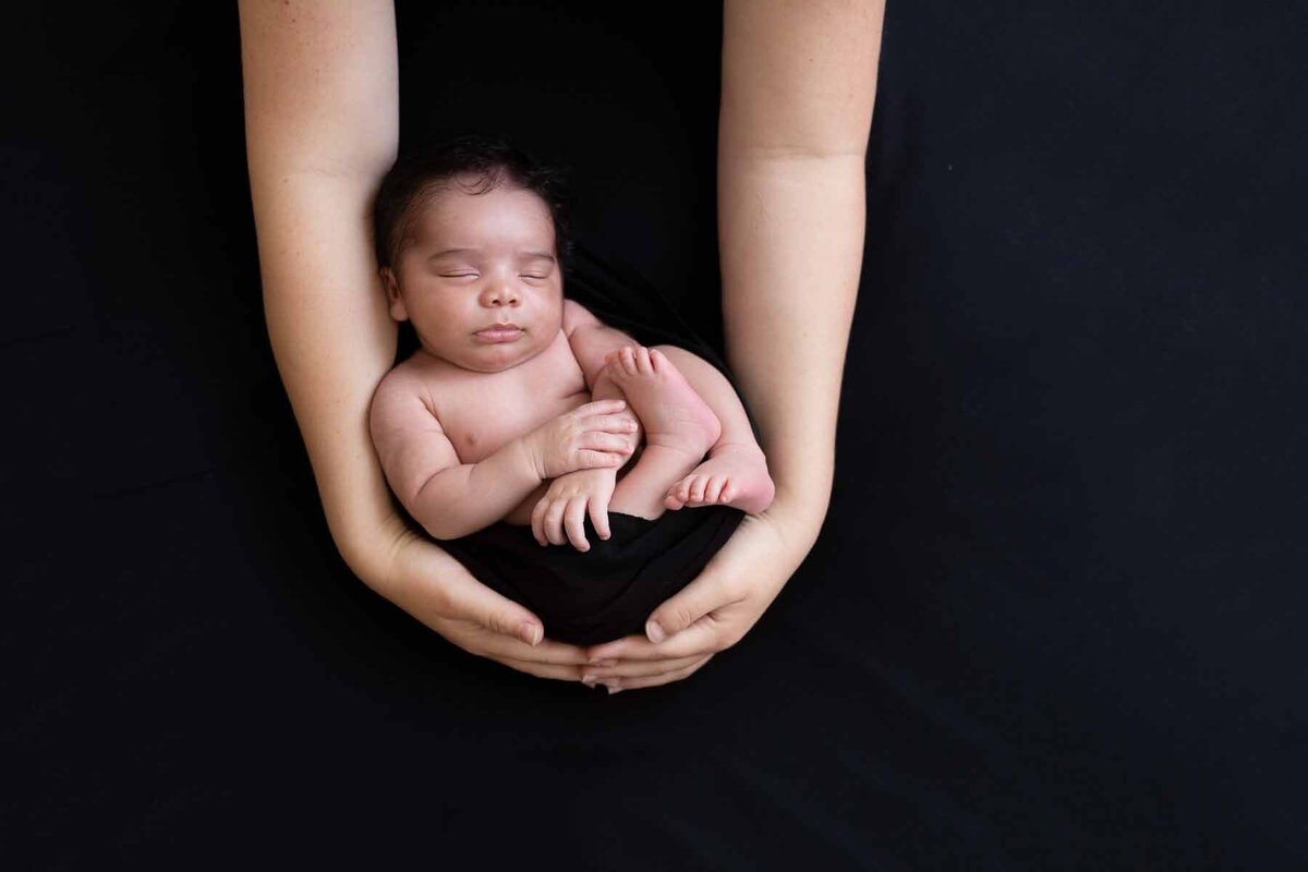 Newborn shoot aan huis op zwarte achtergrond waarbij je baby in moeders handen hangt.