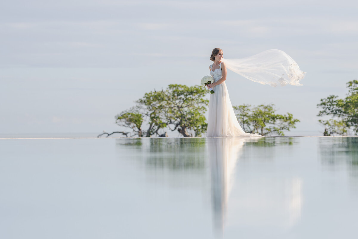 Bride with a flowing veil standing by water, beautifully reflected, captured by Rodrigo Varela Photography.